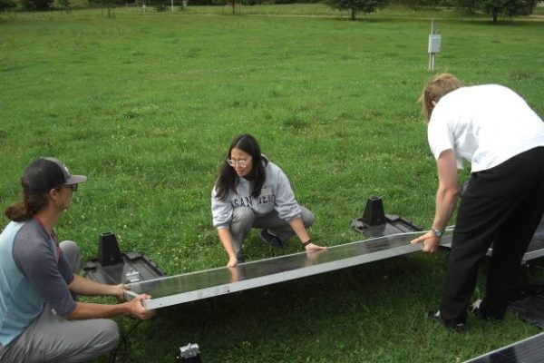 Three young adults are working together in a grassy field to install or move a large, rectangular solar panel. A woman in a grey sweatshirt and glasses crouches in the center, guiding the panel, while two men—one on the left in a baseball cap and one on the right in a white T-shirt—help lift and stabilize it. Several black mounting bases are visible on the grass around them under a bright, overcast sky.