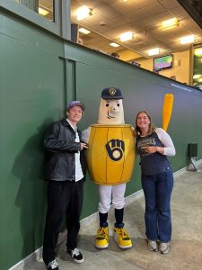 A young man and woman smiling and posing for a photo with the Milwaukee Brewers mascot, Barrelman, inside American Family Field in Milwaukee, WI.