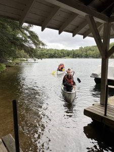 A view from under a rustic wooden boathouse looking out onto a gray, overcast lake. Two silver canoes, each carrying three people wearing orange life jackets, are paddling toward the dock. The shoreline is thick with green pine trees, and the water is clear enough to see the sandy bottom near the pier.