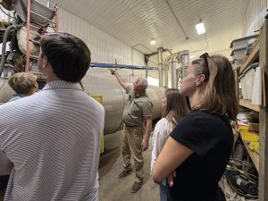 A group of students in a large industrial facility stands around a massive horizontal cylindrical tank. A man in a tan shirt points toward the machinery above the tank, explaining the biodigestion process.
