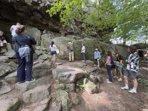 A group of students stands on a rocky, forested trail at Devil's Lake, gathered around a large rock face. An instructor wearing a sun hat and a backpack points toward the rock wall, explaining a geological feature to the group. The students, dressed in casual hiking attire, listen and observe the mossy boulders and overhanging rock formations under the shade of green leafy trees.