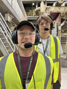Two young men pose for a close-up photo inside a paper mill. They are wearing high-visibility yellow vests, safety glasses, and headsets with microphones. Large industrial machinery is visible in the background.