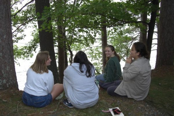 Four young women sit in a circle on a grassy, wooded bank, engaged in conversation. They are framed by large tree trunks and leafy branches, with a bright, shimmering body of water visible in the background. Two of the women are facing the camera and smiling, while the other two are seen from behind. A notebook and phone lie on the ground in the foreground.