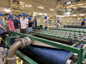 A group of students wearing blue hard hats stands on a metal walkway inside a processing plant. They are observing a large conveyor belt carrying dark organic material, part of the Milorganite production process.