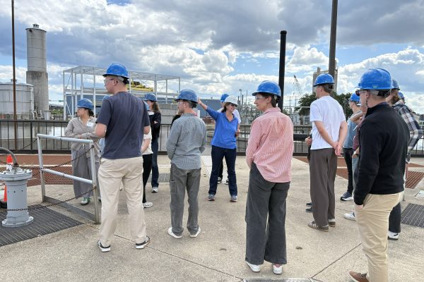 A group of students wearing blue hard hats and safety glasses stand on an outdoor concrete platform during a tour of the Jones Island Water Reclamation Facility. A tour guide in a blue shirt and white hard hat points toward industrial structures, including tanks and a large white metal frame, under a bright sky with dramatic white clouds. The students are dressed in casual clothing and are listening attentively to the presentation.