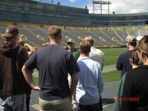 A view from behind a group of students as they stand on the sidelines of Lambeau Field. A tour guide in a Green Bay Packers hat speaks to them, with the vast empty green and yellow stadium seats rising in the background.