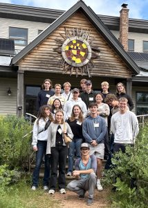 A group of about 20 students and a guide pose for a group photo on the steps of a modern building housing the Midwest Renewable Energy Association. Above them, the wooden gable features a large, colorful sunburst mosaic and solar panels are visible on the roof.
