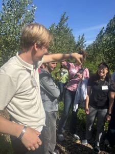 A group of students stands in a sunny field of young trees. One student in the foreground holds up a small, thin garter snake for the others to see, while a female student smiles and looks on.