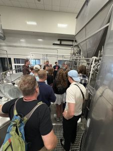 A group of students gathered on a metal industrial mezzanine inside the Titletown Brewery in Green Bay, WI. They are listening to a presentation near large stainless steel vats and complex piping systems.