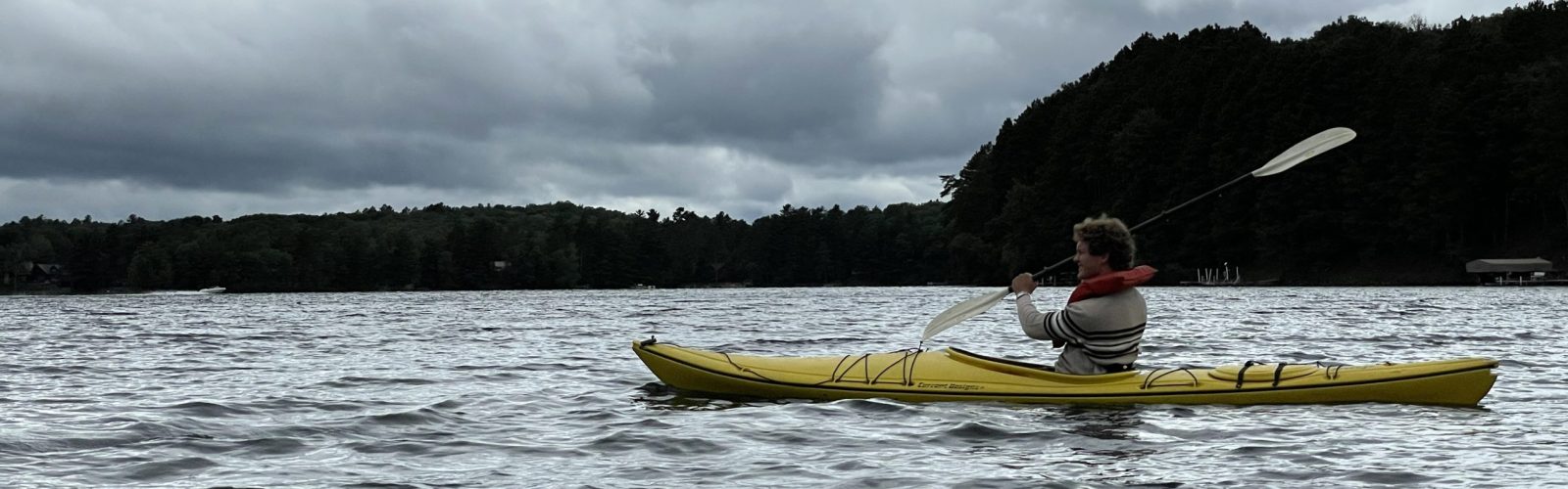 A Madison Torske Klubben student paddling a yellow kayak on a wide lake under a dramatic, cloudy sky. The far shore is lined with dense green trees, and the water has light ripples.