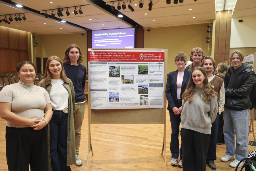 A group of nine smiling young adults—five women and four men—pose in a large, modern auditorium in front of a research poster. The poster, titled "Global Perspectives: Reflections from the SDF Denmark – WI Sustainability Study Tour Fall '25," is part of the Scan Design Fellowship Program at the University of Wisconsin-Madison.